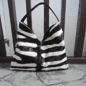 May include: A black and white zebra-striped handbag with a black handle. The bag is made of a textured material and has a wide opening. The bag is sitting on a marble surface.