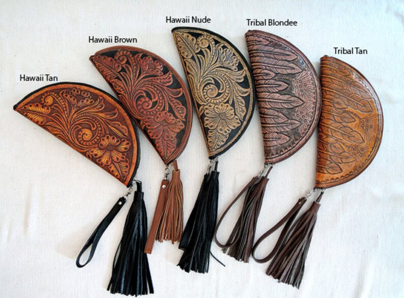 May include: Five half-moon shaped leather clutches with tassel accents. The clutches are decorated with different embossed patterns, including floral designs and feathers. The clutches are labeled "Hawaii Tan", "Hawaii Brown", "Hawaii Nude", "Tribal Blondie", and "Tribal Tan".