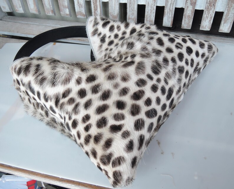 Puede incluir: Un bolso de mano con forma triangular que presenta un estampado animal en blanco y negro con un acabado texturizado similar al pelaje. El bolso incluye una correa negra s&oacute;lida que se curva a lo largo del borde superior, contrastando el dise&ntilde;o.