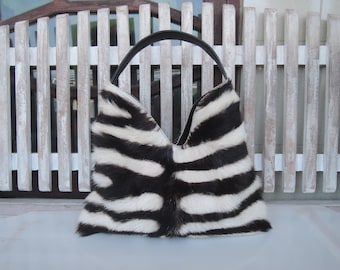 May include: A hobo-style handbag featuring a black and white striped animal print pattern with a textured fur-like finish. The bag has a single black arched handle and a V-shaped top opening, showcasing a distinct contrast between the dark and light tones.
