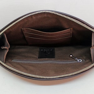 May include: Brown leather clutch with a zipper closure and a small tassel. The clutch has a zippered pocket and a slip pocket inside.