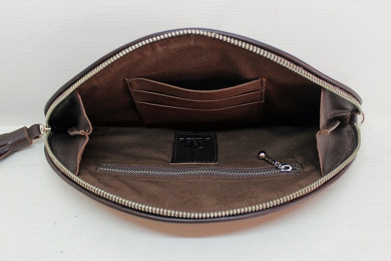 May include: Brown leather clutch with a zipper closure and a small tassel. The clutch has a zippered pocket and a slip pocket inside.