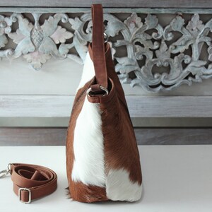May include: A brown and white cowhide leather handbag with a brown leather strap. The bag has a top handle and a detachable shoulder strap.