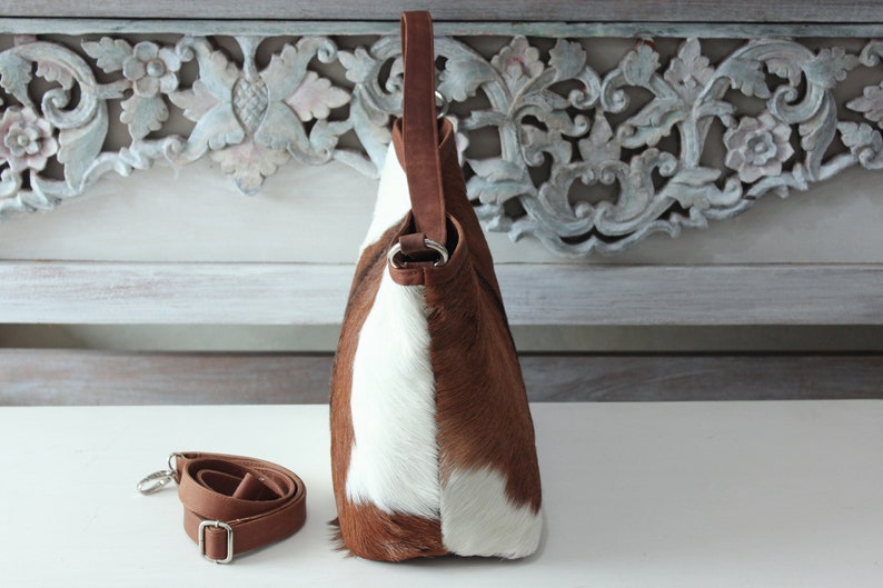May include: A brown and white cowhide leather handbag with a brown leather strap. The bag has a top handle and a detachable shoulder strap.