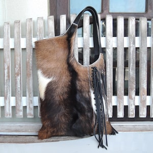 May include: A shoulder bag crafted from tri-coloured cowhide featuring brown, black and white fur patches. The bag includes a black leather handle, a side panel with black lace-up detailing, and long black leather fringe accents hanging from the side.