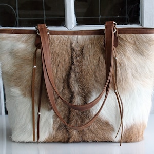 May include: A brown and white cowhide tote bag with brown leather straps and tassels. The bag has a top zipper closure and a spacious interior.