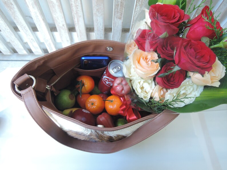 May include: A brown leather tote bag with a white fur lining. The bag is filled with fruit, a can of soda, a cell phone, and a bouquet of red and white roses.