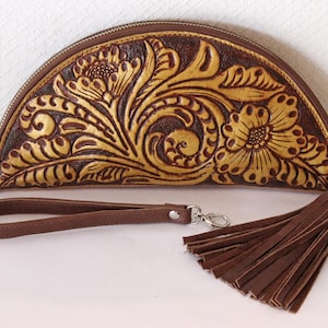 Pode incluir: Uma carteira de mão de couro em forma de meia-lua com um design floral em dourado e castanho. A carteira de mão tem um fecho de correr e uma alça de couro castanho com um tassel.