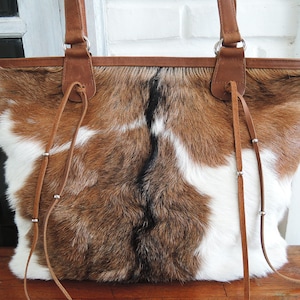 May include: A brown, white, and black cowhide tote bag with brown leather handles and trim. The bag has a rectangular shape and features long, thin leather straps with silver accents. The bag is sitting on a wooden surface.