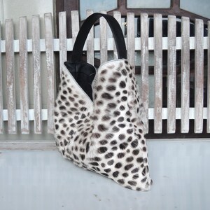 Puede incluir: Un bolso de estilo hobo con un estampado de manchas blancas y negras sobre una superficie texturizada. El bolso tiene una sola asa negra y una abertura profunda en forma de V que revela un forro interior negro y liso.