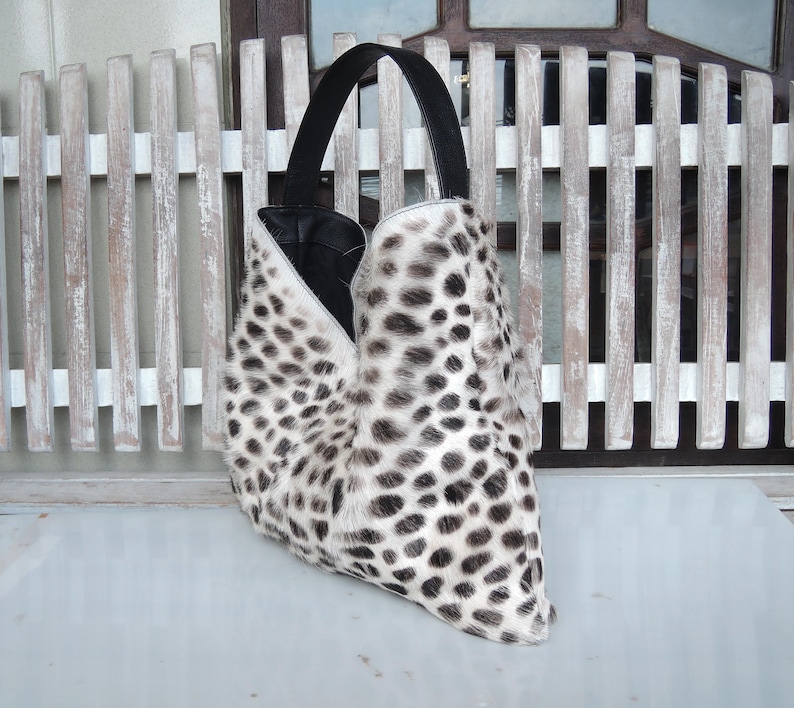 Puede incluir: Un bolso de estilo hobo con un estampado de manchas blancas y negras sobre una superficie texturizada. El bolso tiene una sola asa negra y una abertura profunda en forma de V que revela un forro interior negro y liso.