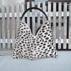 Puede incluir: Un bolso de mano estilo hobo con un patr&oacute;n de estampado animal en blanco y negro con una apariencia texturizada similar al pelaje. El bolso tiene una abertura superior en forma de V profunda y un asa negra &uacute;nica en los bordes superiores.