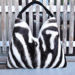 Könnte beinhalten: Eine Handtasche im Hobo-Stil mit schwarz-weißem Zebramuster und einer texturierten Oberfläche aus Fell. Die Tasche hat eine tiefe V-förmige Öffnung an der Oberseite und einen einzelnen, festen schwarzen Ledergriff an der oberen Mitte.