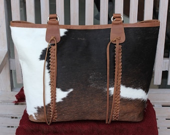 Puede incluir: Un bolso tote con estampado de piel de vaca con parches de pelo negro, marrón y blanco. El bolso incluye un borde de cuero marrón en la parte superior y dos asas de cuero marrón con detalles trenzados que se extienden por la parte frontal.