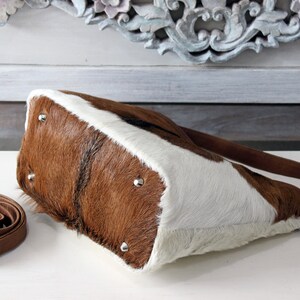 May include: A brown and white cowhide tote bag with a brown leather strap. The bag has a flap closure and a silver buckle.
