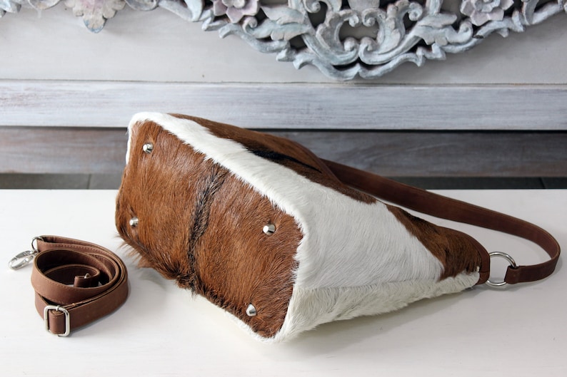 May include: A brown and white cowhide tote bag with a brown leather strap. The bag has a flap closure and a silver buckle.