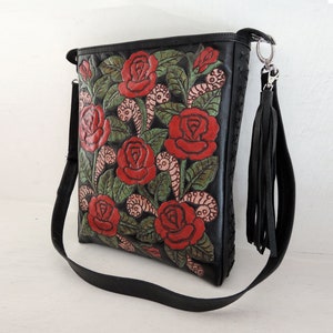 May include: Black leather shoulder bag with a floral design featuring red roses and green leaves. The bag has a long adjustable strap and a tassel.