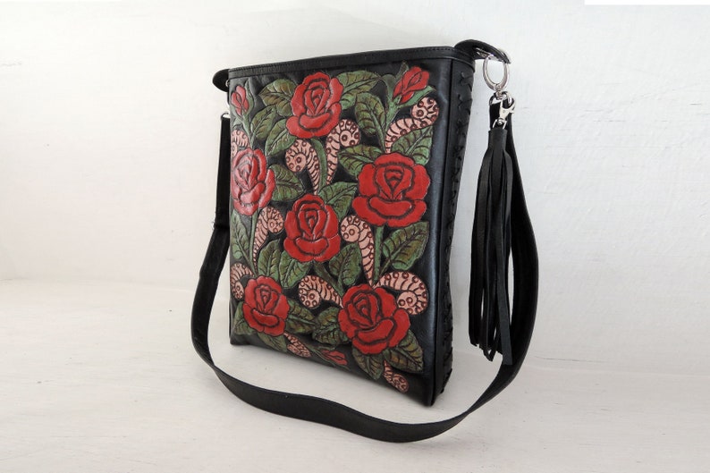 May include: Black leather shoulder bag with a floral design featuring red roses and green leaves. The bag has a long adjustable strap and a tassel.