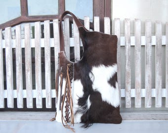 May include: A hobo-style shoulder bag crafted from brown and white cowhide with a textured fur finish. The bag features a single brown leather strap, a side zipper closure, and decorative leather fringe accents with a braided lace-up detail.
