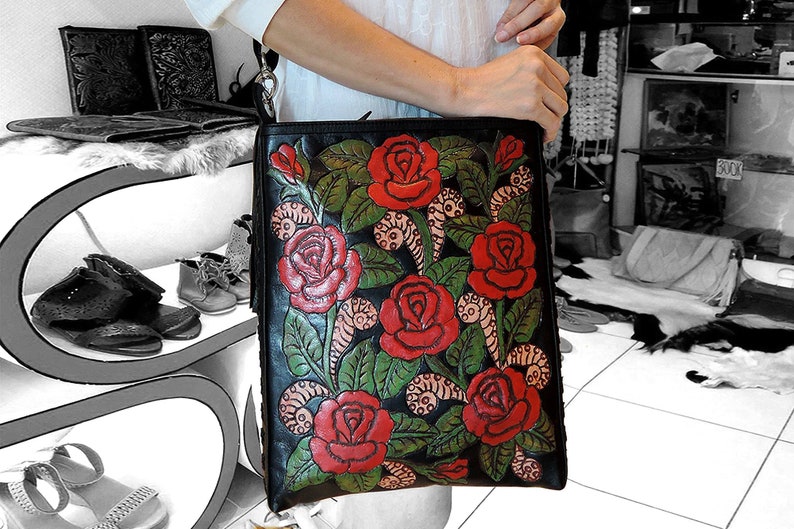 May include: A black leather tote bag with a floral design. The bag is decorated with red roses and green leaves. The bag has a long strap and a zip closure.