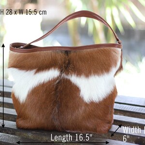 May include: A brown and white cowhide tote bag with a brown leather handle. The bag measures 16.5 inches long, 11 inches high, and 6 inches wide at the base.