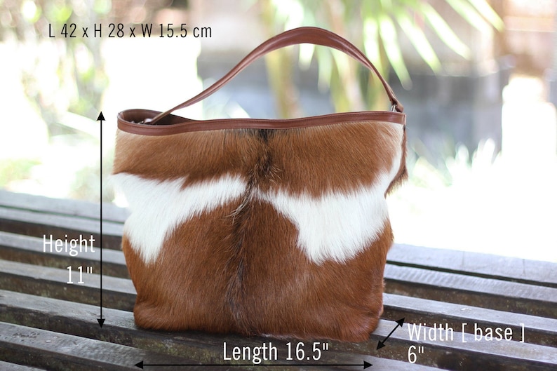 May include: A brown and white cowhide tote bag with a brown leather handle. The bag measures 16.5 inches long, 11 inches high, and 6 inches wide at the base.