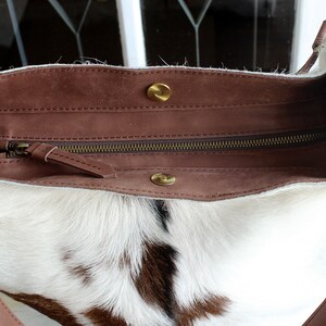 Puede incluir: Un bolso tote de piel de vaca blanca y marr&oacute;n con una solapa de cuero marr&oacute;n y cierre de cremallera. El bolso tiene dos anillos met&aacute;licos dorados en las asas.