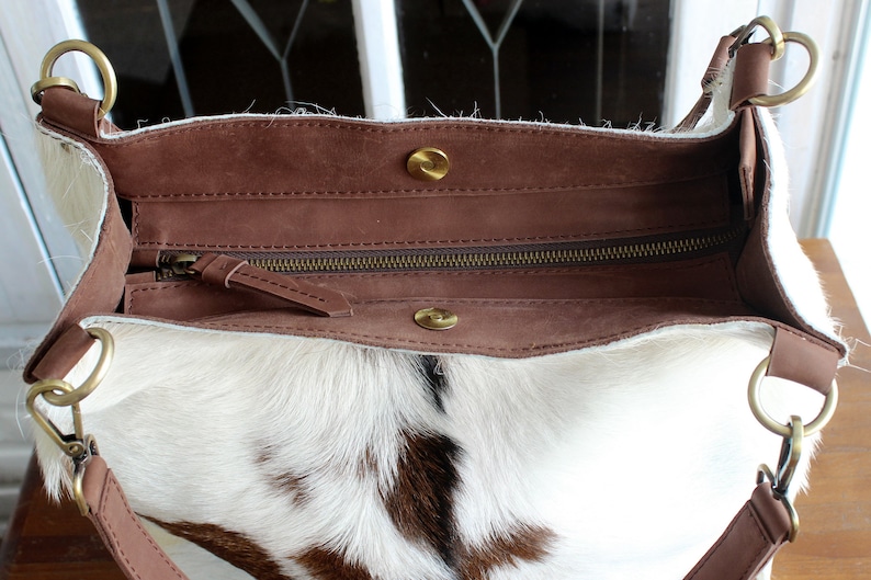 Puede incluir: Un bolso tote de piel de vaca blanca y marr&oacute;n con una solapa de cuero marr&oacute;n y cierre de cremallera. El bolso tiene dos anillos met&aacute;licos dorados en las asas.