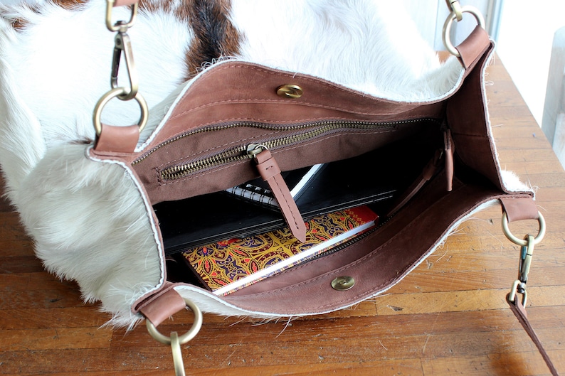 Puede incluir: Un bolso tote de piel de vaca blanca y marr&oacute;n con una parte superior de cuero marr&oacute;n con cremallera y una correa de cuero marr&oacute;n. El bolso est&aacute; abierto y tiene varios cuadernos y un libro en su interior.