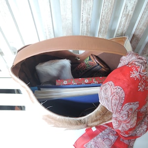 Puede incluir: Un bolso hobo con asa de cuero color canela y exterior de piel de vaca marr&oacute;n y blanca. El interior contiene una pila de papeles, un cuaderno rojo con estampado floral, una peque&ntilde;a bolsa de piel blanca y una cartera oscura con estampado floral.