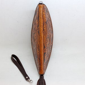 May include: A brown leather clutch with a floral design and a tassel. The clutch has a brown leather strap with a silver clasp.