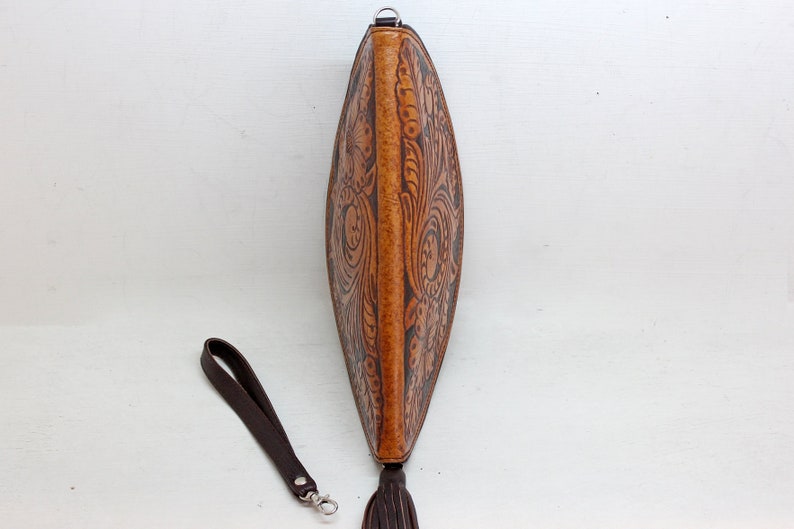 May include: A brown leather clutch with a floral design and a tassel. The clutch has a brown leather strap with a silver clasp.