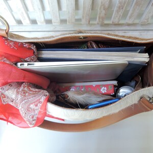 Puede incluir: Un bolso tote de cuero marr&oacute;n con ribete de piel blanca. El bolso est&aacute; abierto y lleno de art&iacute;culos, incluyendo un port&aacute;til, cuadernos, una bufanda roja y una pluma.