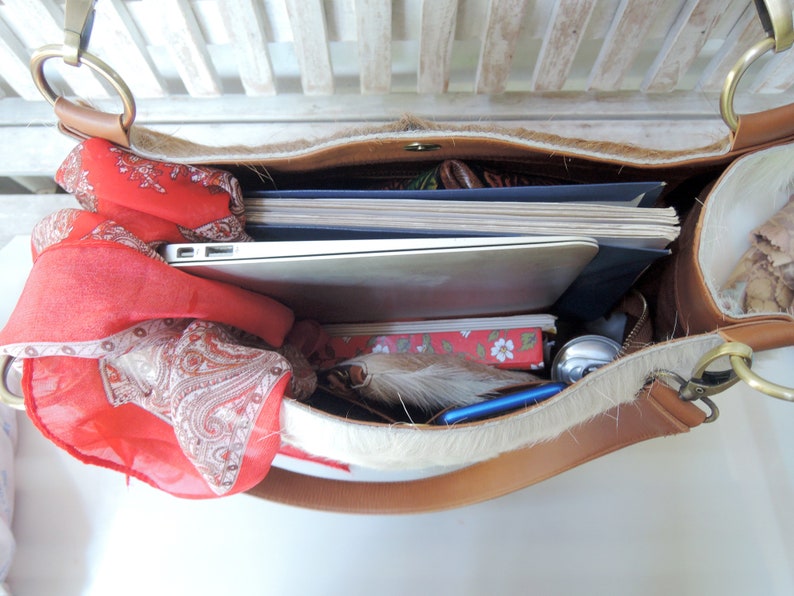 Puede incluir: Un bolso tote de cuero marr&oacute;n con ribete de piel blanca. El bolso est&aacute; abierto y lleno de art&iacute;culos, incluyendo un port&aacute;til, cuadernos, una bufanda roja y una pluma.