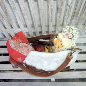 Puede incluir: Un bolso tote de piel de vaca blanca y marr&oacute;n con una larga correa de cuero marr&oacute;n. El bolso est&aacute; lleno de un ramo de rosas blancas y melocot&oacute;n y una bufanda estampada en rojo y blanco.