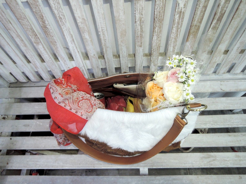Puede incluir: Un bolso tote de piel de vaca blanca y marr&oacute;n con una larga correa de cuero marr&oacute;n. El bolso est&aacute; lleno de un ramo de rosas blancas y melocot&oacute;n y una bufanda estampada en rojo y blanco.