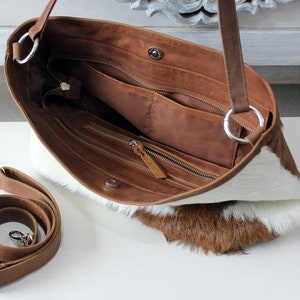 May include: A brown leather handbag with a white and brown cowhide exterior. The bag has a zippered closure and an adjustable shoulder strap. The bag is open and the interior is visible, showing a brown lining and multiple pockets.