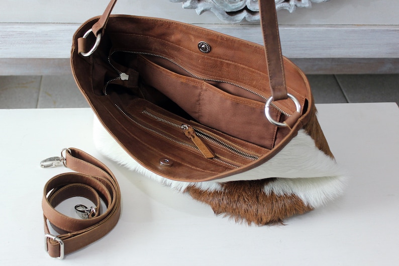 May include: A brown leather handbag with a white and brown cowhide exterior. The bag has a zippered closure and an adjustable shoulder strap. The bag is open and the interior is visible, showing a brown lining and multiple pockets.