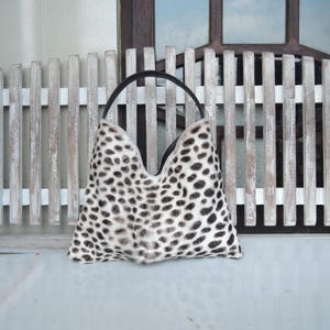 Puede incluir: Un bolso de mano estilo hobo con un estampado animal en blanco y negro y una textura similar al pelaje. El bolso presenta una abertura superior en forma de V profunda y una &uacute;nica asa negra sujeta a los bordes superiores, formando una silueta triangular.