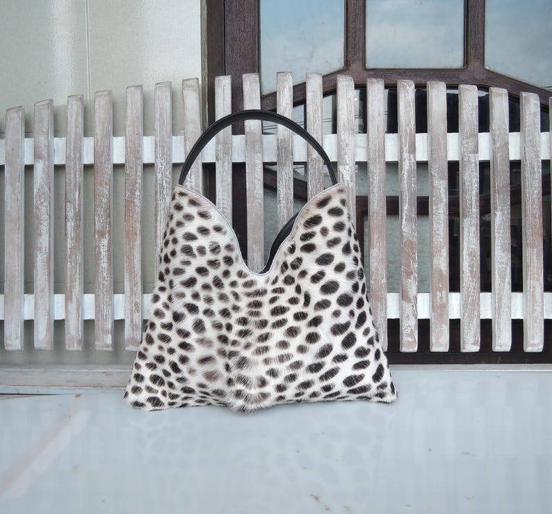 Puede incluir: Un bolso de mano estilo hobo con un estampado animal en blanco y negro y una textura similar al pelaje. El bolso presenta una abertura superior en forma de V profunda y una &uacute;nica asa negra sujeta a los bordes superiores, formando una silueta triangular.