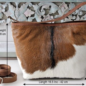 May include: Brown and white cowhide tote bag with a brown leather strap. The bag measures 11 inches to 28 inches in height, 16.5 inches to 42 inches in length, and 6 inches to 15.5 inches in width.