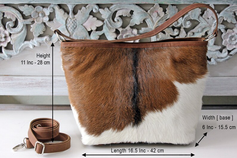 May include: Brown and white cowhide tote bag with a brown leather strap. The bag measures 11 inches to 28 inches in height, 16.5 inches to 42 inches in length, and 6 inches to 15.5 inches in width.