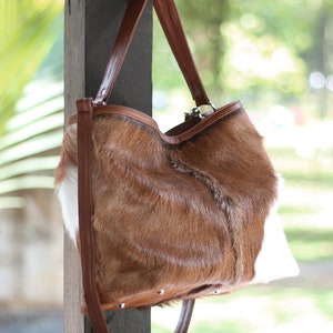 May include: A brown and white cowhide tote bag with a brown leather strap. The bag is hanging on a wooden post.