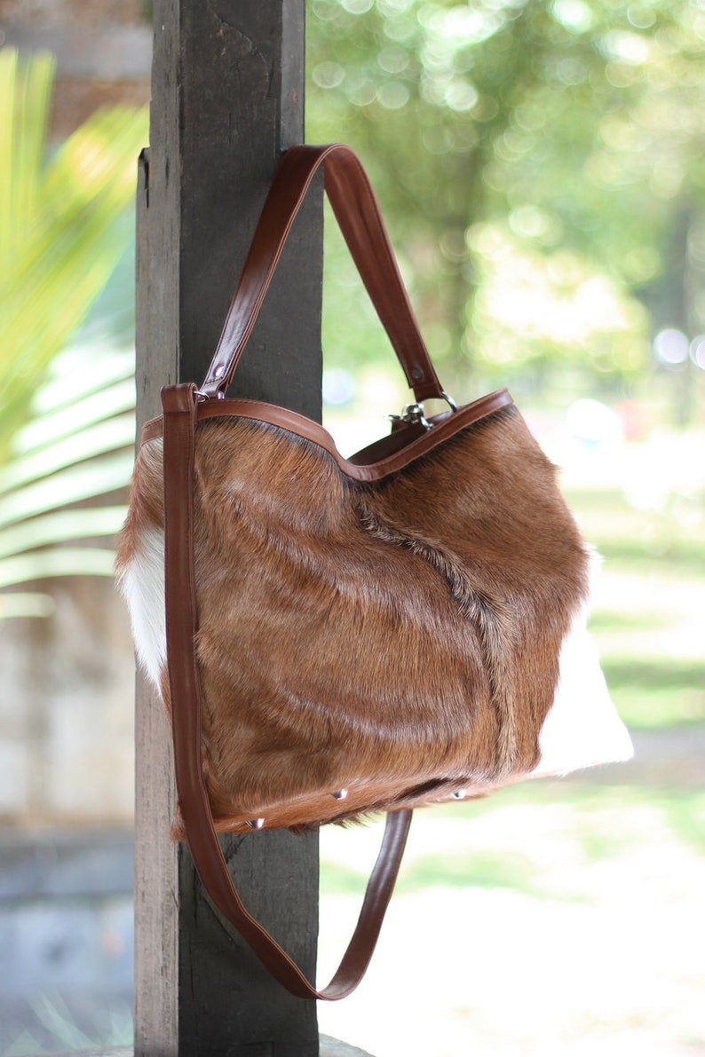 May include: A brown and white cowhide tote bag with a brown leather strap. The bag is hanging on a wooden post.
