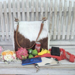 Puede incluir: Un bolso tote de piel de vaca blanca y marr&oacute;n con una correa de cuero marr&oacute;n, una lata de Coca-Cola, un fruto del drag&oacute;n, limas, pl&aacute;tanos, una cartera negra, una bufanda roja y un ramo de rosas rosas y blancas en un banco de madera blanco.