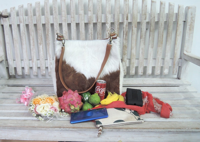 Puede incluir: Un bolso tote de piel de vaca blanca y marr&oacute;n con una correa de cuero marr&oacute;n, una lata de Coca-Cola, un fruto del drag&oacute;n, limas, pl&aacute;tanos, una cartera negra, una bufanda roja y un ramo de rosas rosas y blancas en un banco de madera blanco.