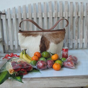 May include: A brown and white cowhide handbag with a brown leather handle, a bouquet of red roses, a can of Coca-Cola, and a variety of fruits including limes, oranges, and bananas.