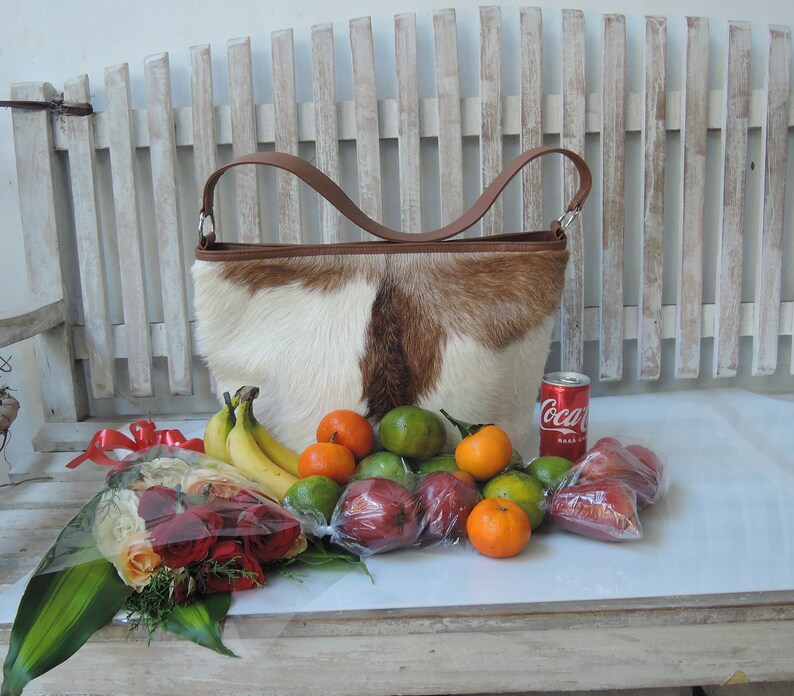 May include: A brown and white cowhide handbag with a brown leather handle, a bouquet of red roses, a can of Coca-Cola, and a variety of fruits including limes, oranges, and bananas.