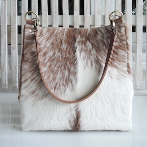 May include: A large, rectangular cowhide tote bag with a brown and white spotted pattern. The bag has brown leather straps and gold-colored metal rings. The bag is sitting in front of a white wooden fence.