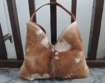 Puede incluir: Un bolso de estilo hobo confeccionado en piel de vaca marrón y blanca con un acabado texturizado y peludo. El bolso cuenta con una sola correa de hombro de cuero color canela y una abertura superior en forma de V con un patrón moteado.